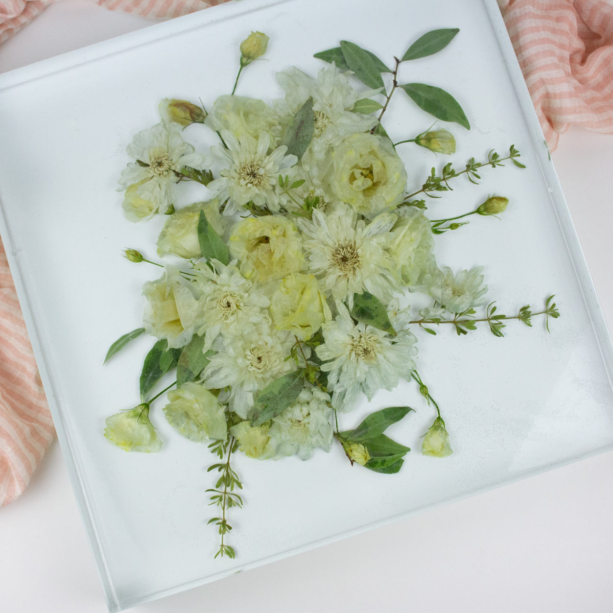 A customary floral arrangement with a variety of dried flowers in shades of yellow and green, displayed in a clear square block on a white surface, with a striped fabric in the background.