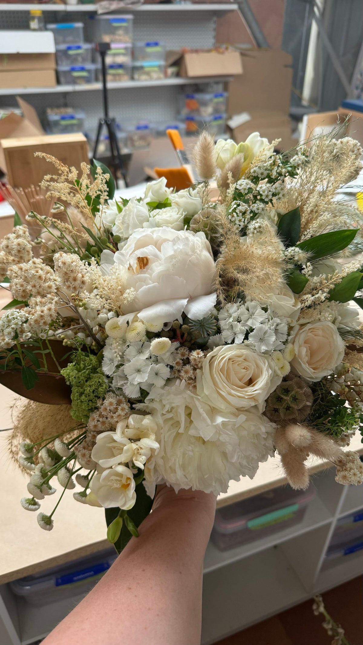 A hand holding a large, rustic dried flower bouquet featuring cream and white roses, baby's breath, pampas grass, and green foliage in a workshop setting.