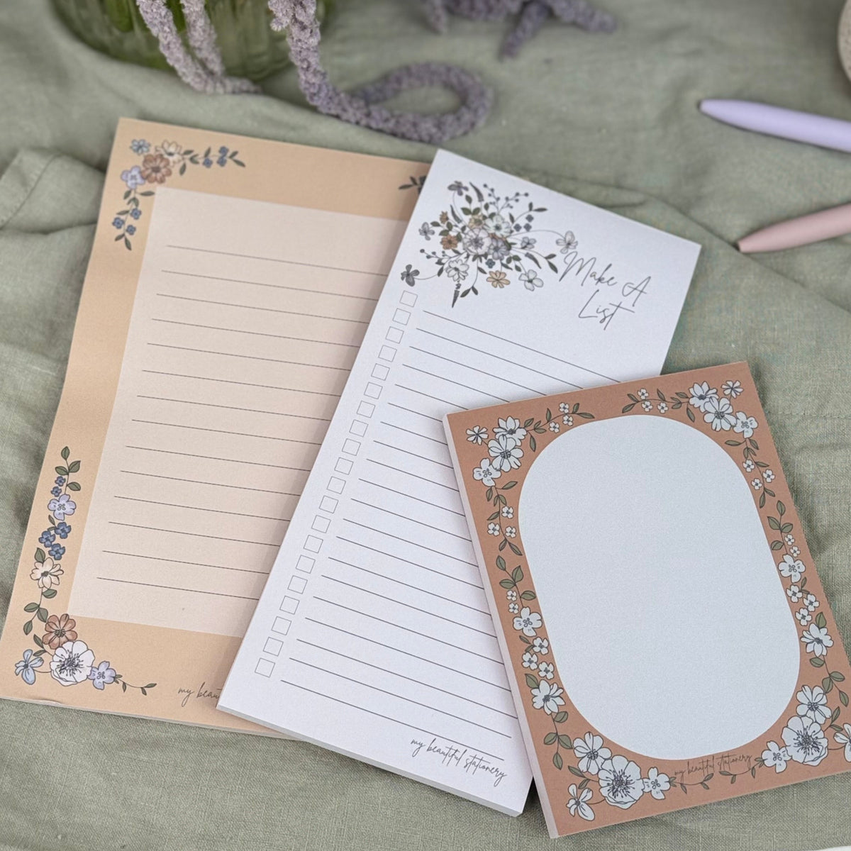 Set of three decorative notepads on a green surface with flowers and a pen.