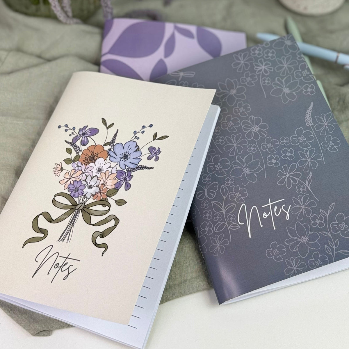 Two notebooks with floral designs on a surface with flowers and a pen holder in the background.