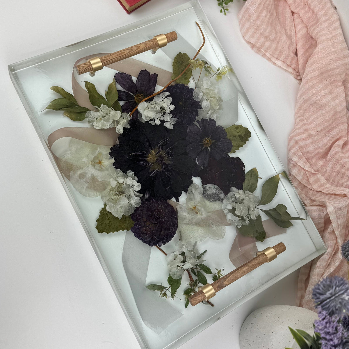 Decorative tray with pressed flowers, books, and a pink cloth on a white surface.