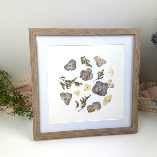 A square framed pressed flower art print with a light oak wood frame and white mat, featuring dried lavender and yellow flowers on a white background, styled on a white surface with soft pink fabric and greenery.