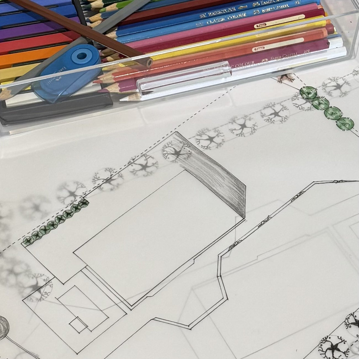 A close-up of a hand-drawn architectural floor plan or design sketch on white paper, with colored pencils and drawing tools arranged in a clear container above it.