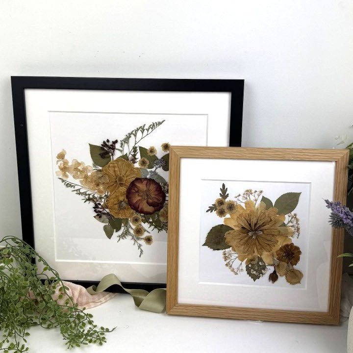 Two framed pressed flower art prints displayed on a white surface against a white wall. One print has a black frame and the other has a light wood frame, both with white mats and floral arrangements in muted tan, brown, olive green, and cream tones.